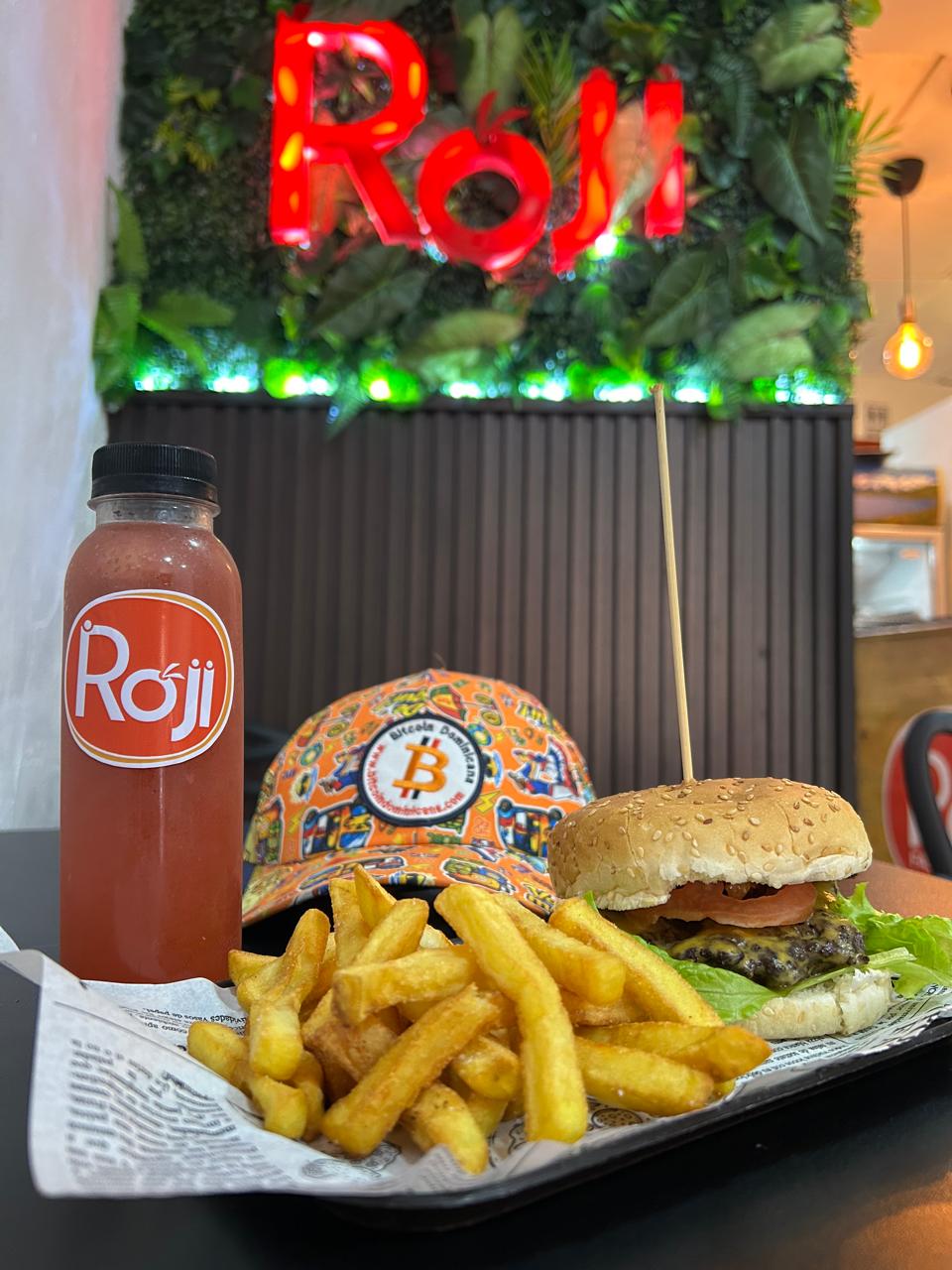 Hamburguesa, papas fritas y jugo Roji en bandeja junto a gorra de Bitcoin Dominicana, con letrero neón Rojis al fondo