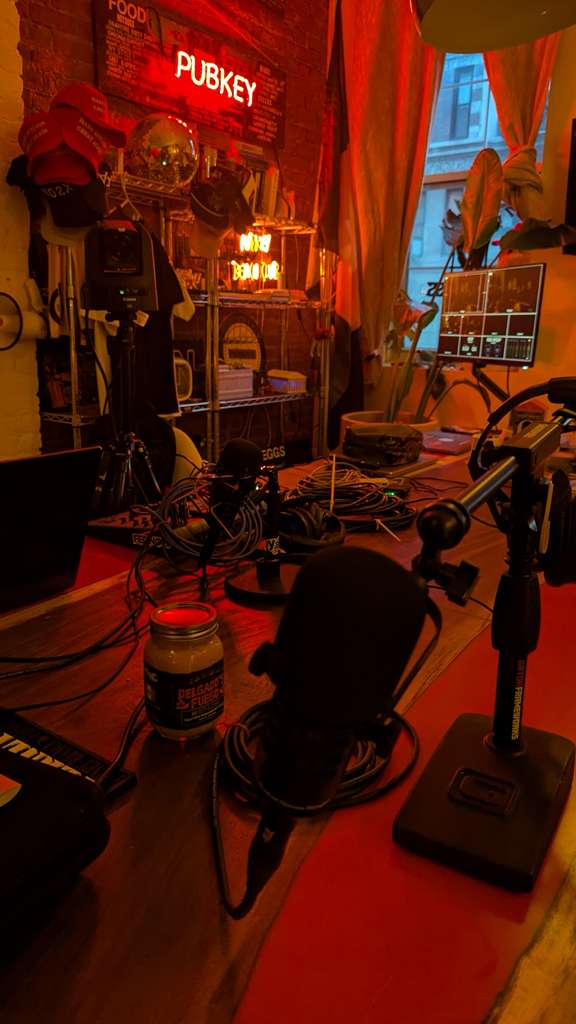Set de grabación de podcast con micrófonos y luces rojas bajo el letrero de neón PUBKEY
