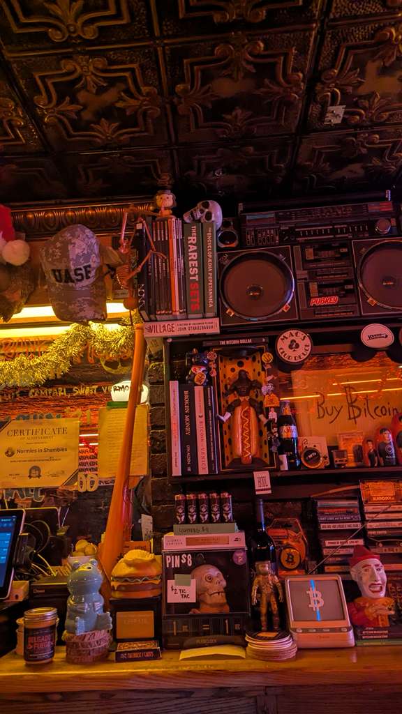 Estantería con libros, memorabilia y figuras de Bitcoin junto a un cartel que dice Buy Bitcoin en PubKey