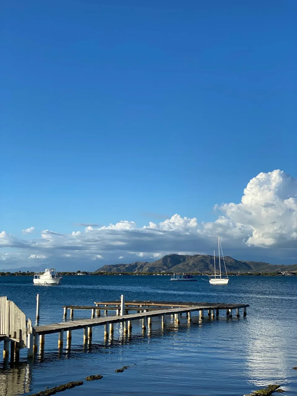 Un muelle de madera se adentra en aguas tranquilas de una bahía, con veleros anclados y montañas al fondo, en la costa dominicana.