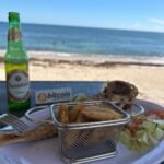 Plato de pescado frito con tostones en una canasta metálica, una cerveza Presidente y un pequeño letrero con el logo de Bitcoin, sobre una mesa frente al mar en una playa dominicana.