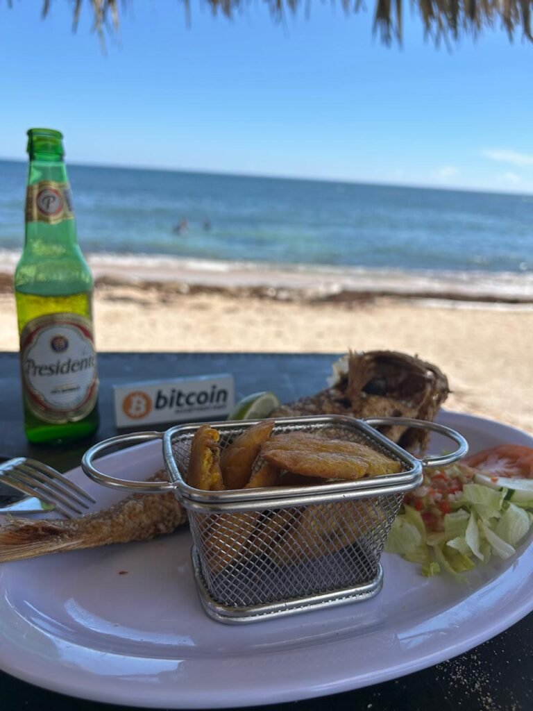 Plato de pescado frito con tostones en una canasta metálica, una cerveza Presidente y un pequeño letrero con el logo de Bitcoin, sobre una mesa frente al mar en una playa dominicana.