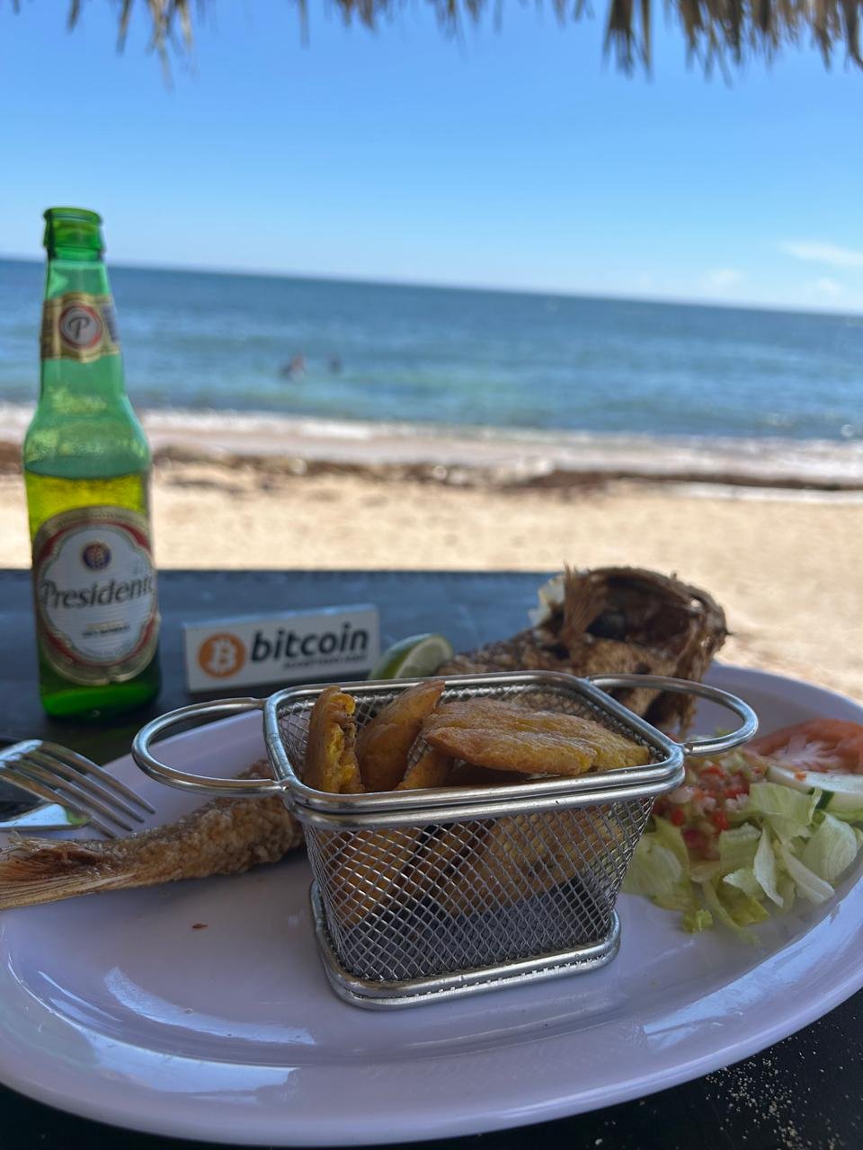 Plato de pescado frito con tostones en una canasta metálica, una cerveza Presidente y un pequeño letrero con el logo de Bitcoin, sobre una mesa frente al mar en una playa dominicana.