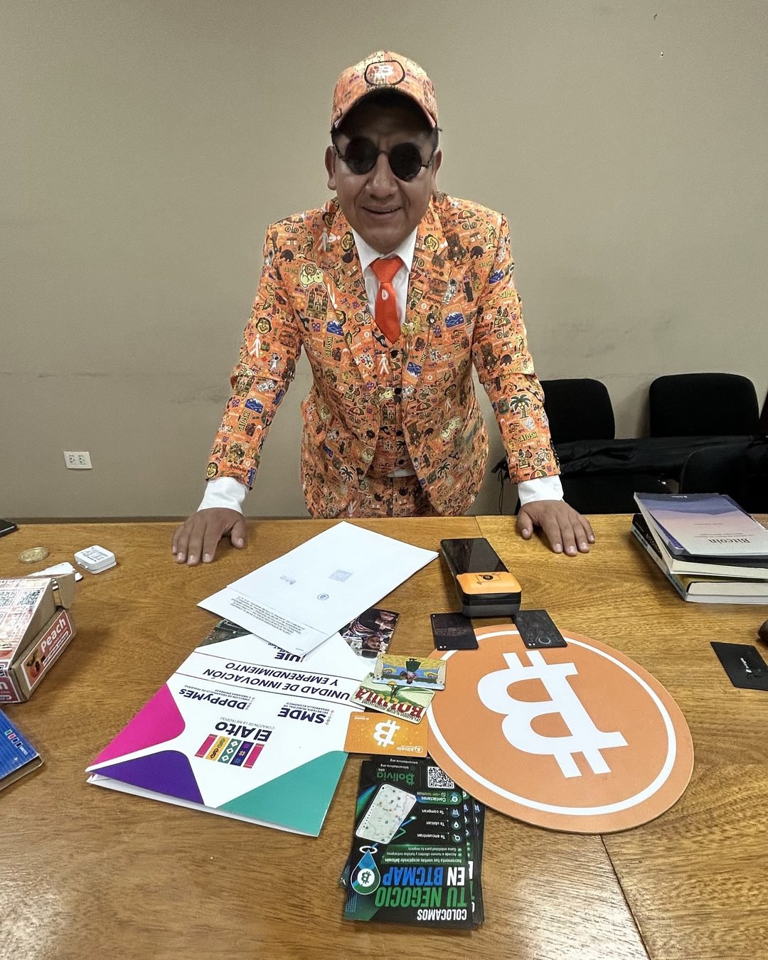 Achachilabtc, con traje y gorra naranjas, apoyado en una mesa con material de Bitcoin y del municipio de El Alto
