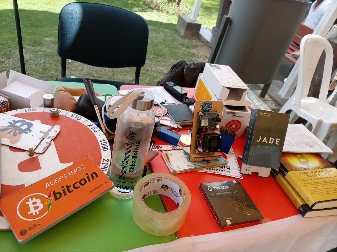 Mesa de Bitcoin Bolivia con un cartel «Aceptamos bitcoin», un nodo, una billetera Jade, una Trezor y libros sobre Bitcoin, en el AWS Student Community Day de Cochabamba