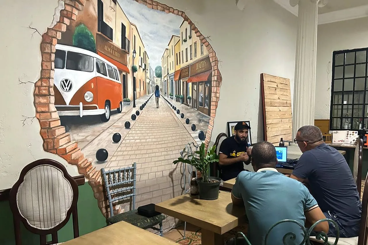Carlos Gutiérrez, de Quisqueya Project, explica Bitcoin para principiantes en una mesa con laptop, frente a un mural de una calle colonial en The Perpetual Lab