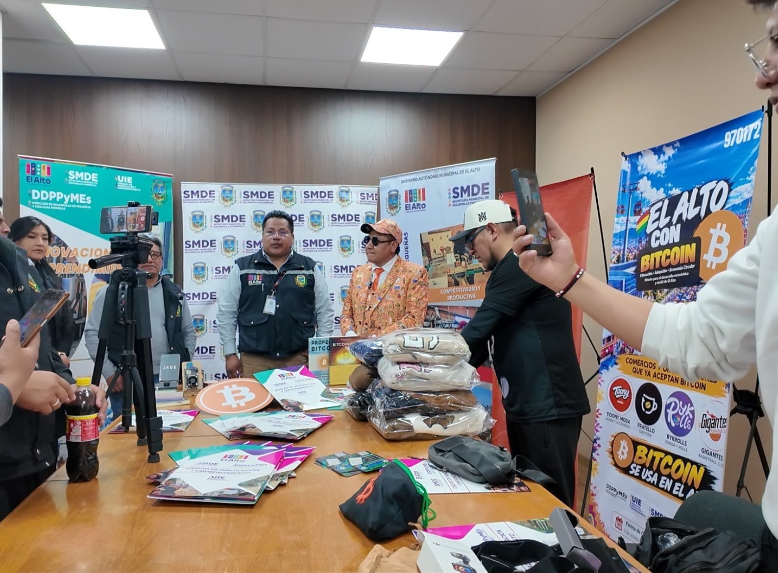 Achachilabtc y un funcionario municipal frente a una mesa con material de Bitcoin, junto a banners de la Secretaría Municipal de Desarrollo Económico y el cartel «El Alto con Bitcoin» con los comercios que ya aceptan bitcoin