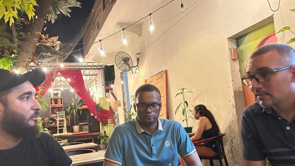 Carlos Gutiérrez, de Quisqueya Project (izquierda), conversa sobre Bitcoin con dos participantes en el patio de The Perpetual Lab, en la Zona Colonial de Santo Domingo, de noche y bajo luces colgantes