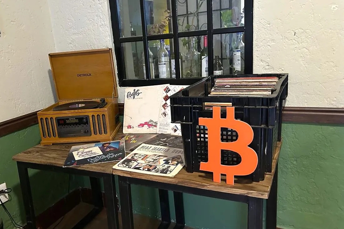 Mesa con un tocadiscos, discos de vinilo y un símbolo naranja de Bitcoin en The Perpetual Lab